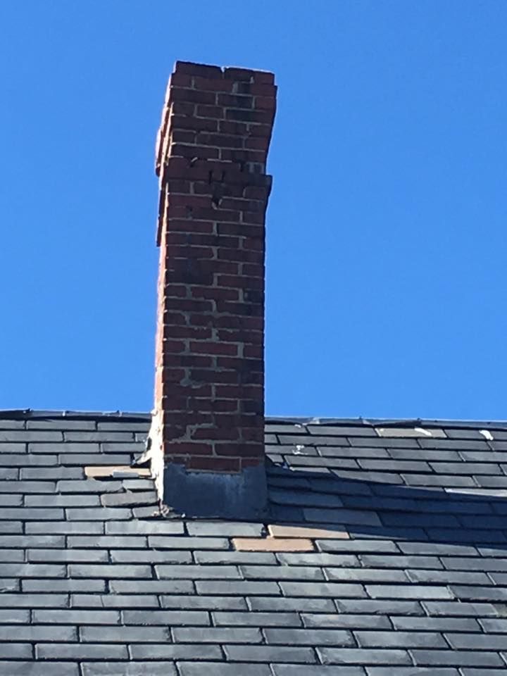 Brick chimney on a dark shingle roof against a clear blue sky. The top is damaged.