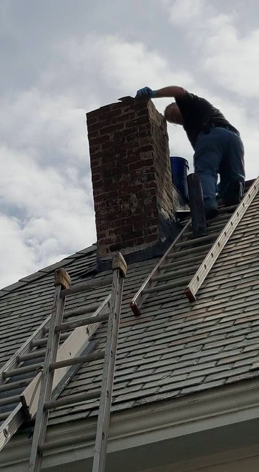 Man repairing a brick chimney on a rooftop, using two ladders. Cloudy sky.