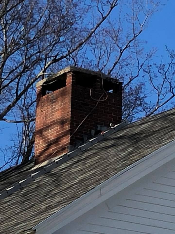 Brick chimney on a house roof against a blue sky, bare tree branches in background.