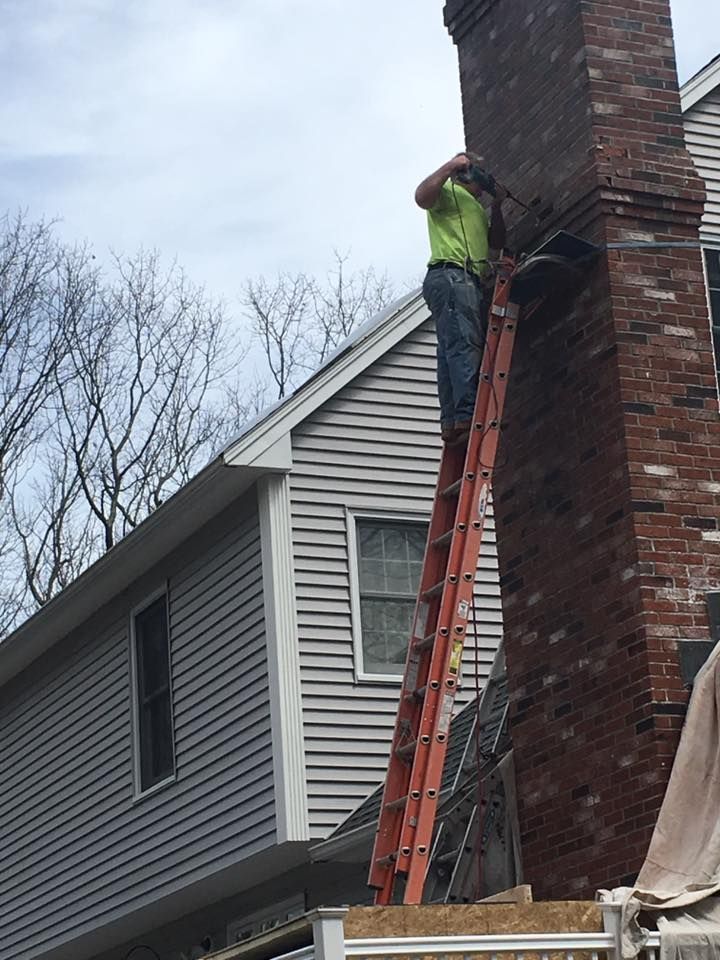 Person on a ladder repairing a brick chimney on a house with gray siding.