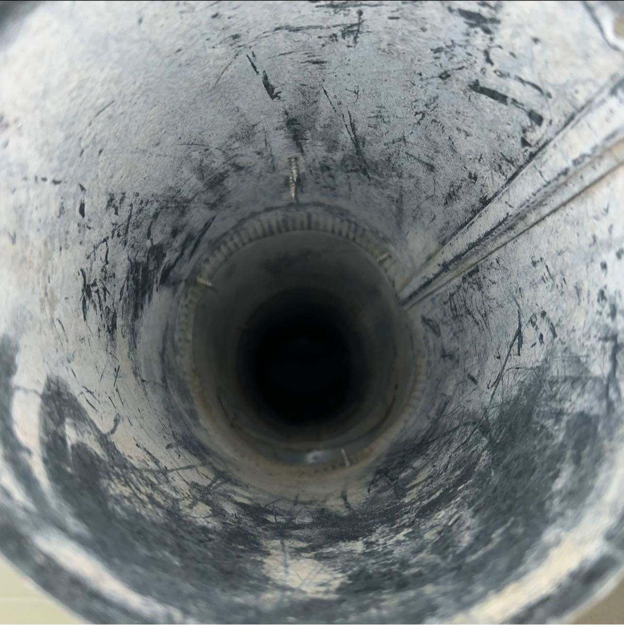Looking down the inside of a metal pipe, with a dark opening at the bottom and scratches on the walls.