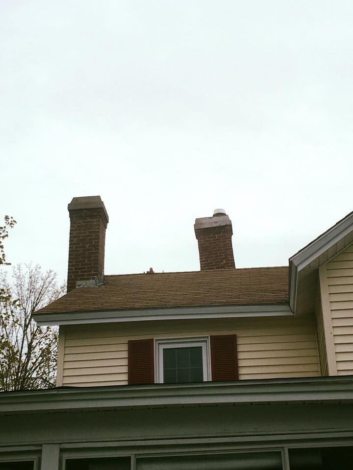Two brick chimneys on a house roof with red shutters and yellow siding against a cloudy sky.