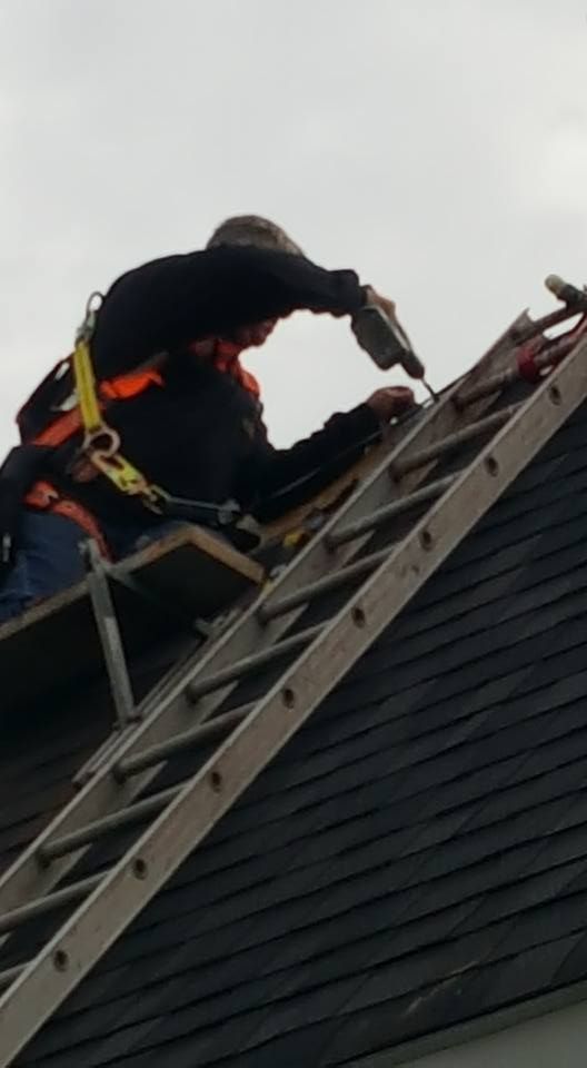 Roofer using a drill on a dark shingle roof, secured by a harness. Ladder angled towards the roof.