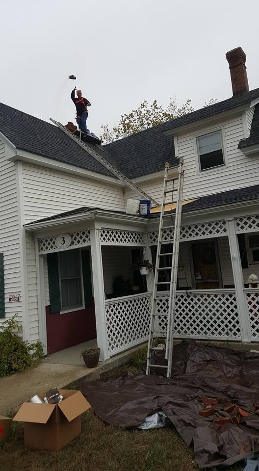 Man on roof working, tall ladder propped up. House with white siding, porch. Cloudy day.