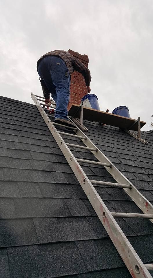 A person on a ladder works on a brick chimney on a rooftop under a cloudy sky.