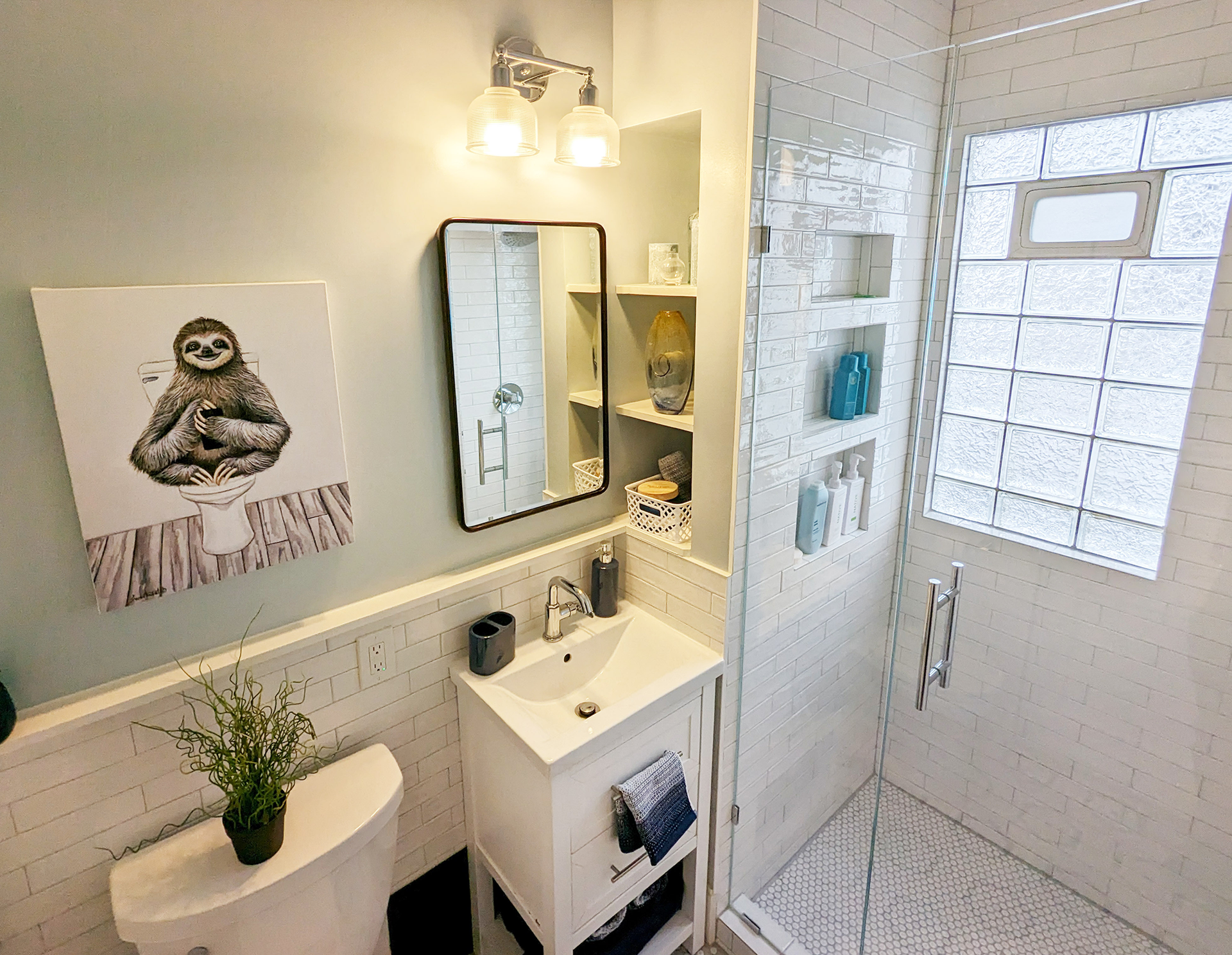 A light-colored bathroom featuring a small white pedestal sink, a wall-mounted mirror, a sloth print, and a glass shower.