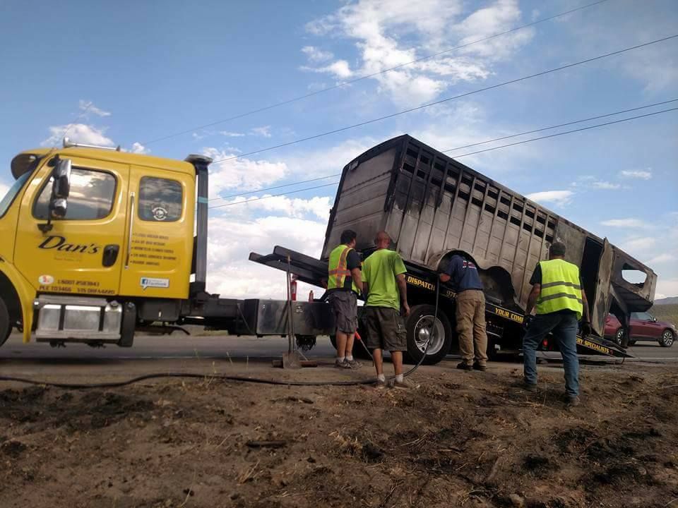 Yellow tow truck lifts a partially burned livestock trailer; people in vests assess damage roadside.