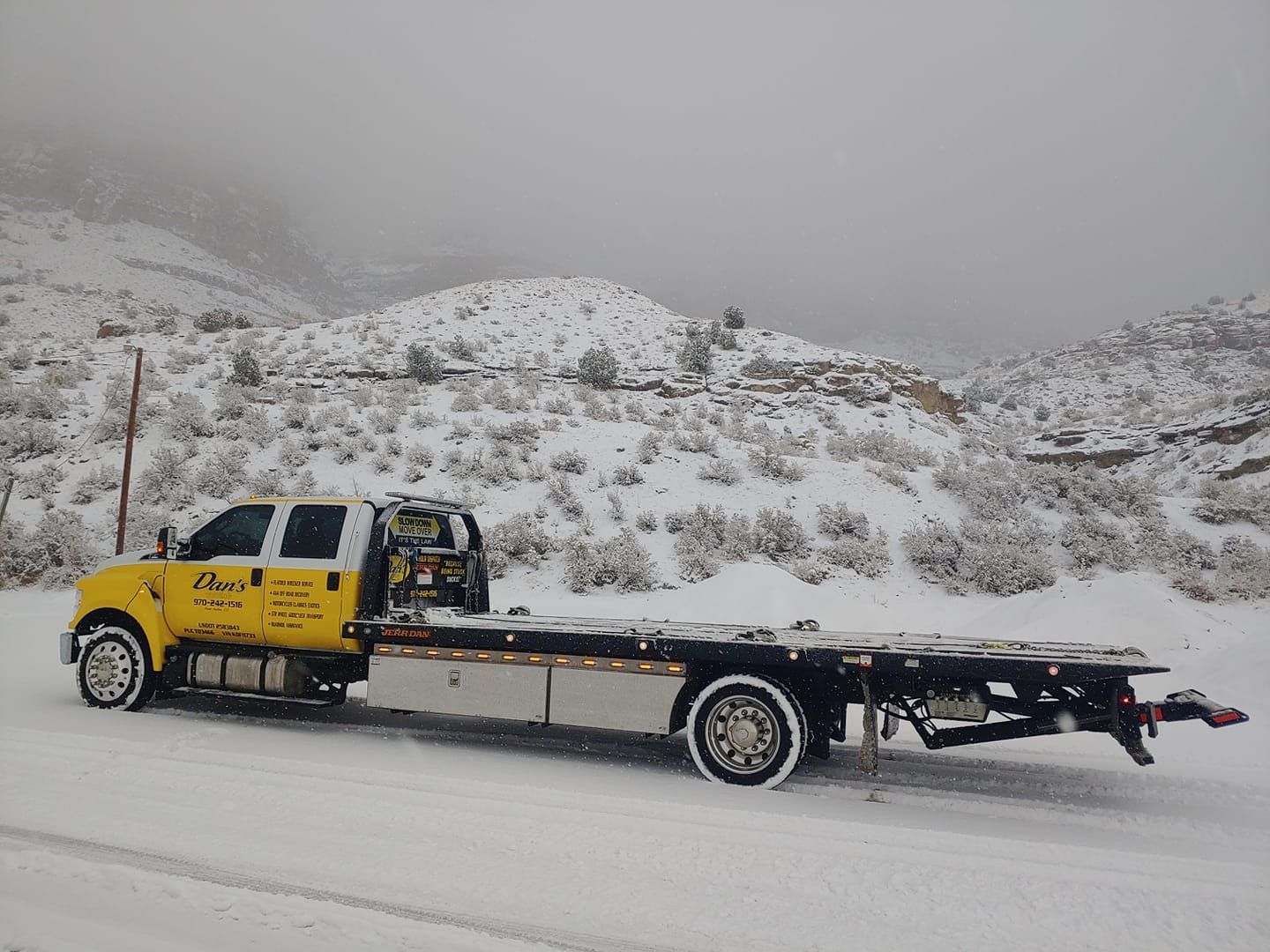 Yellow tow truck on a snowy road in front of a snow-covered hill.
