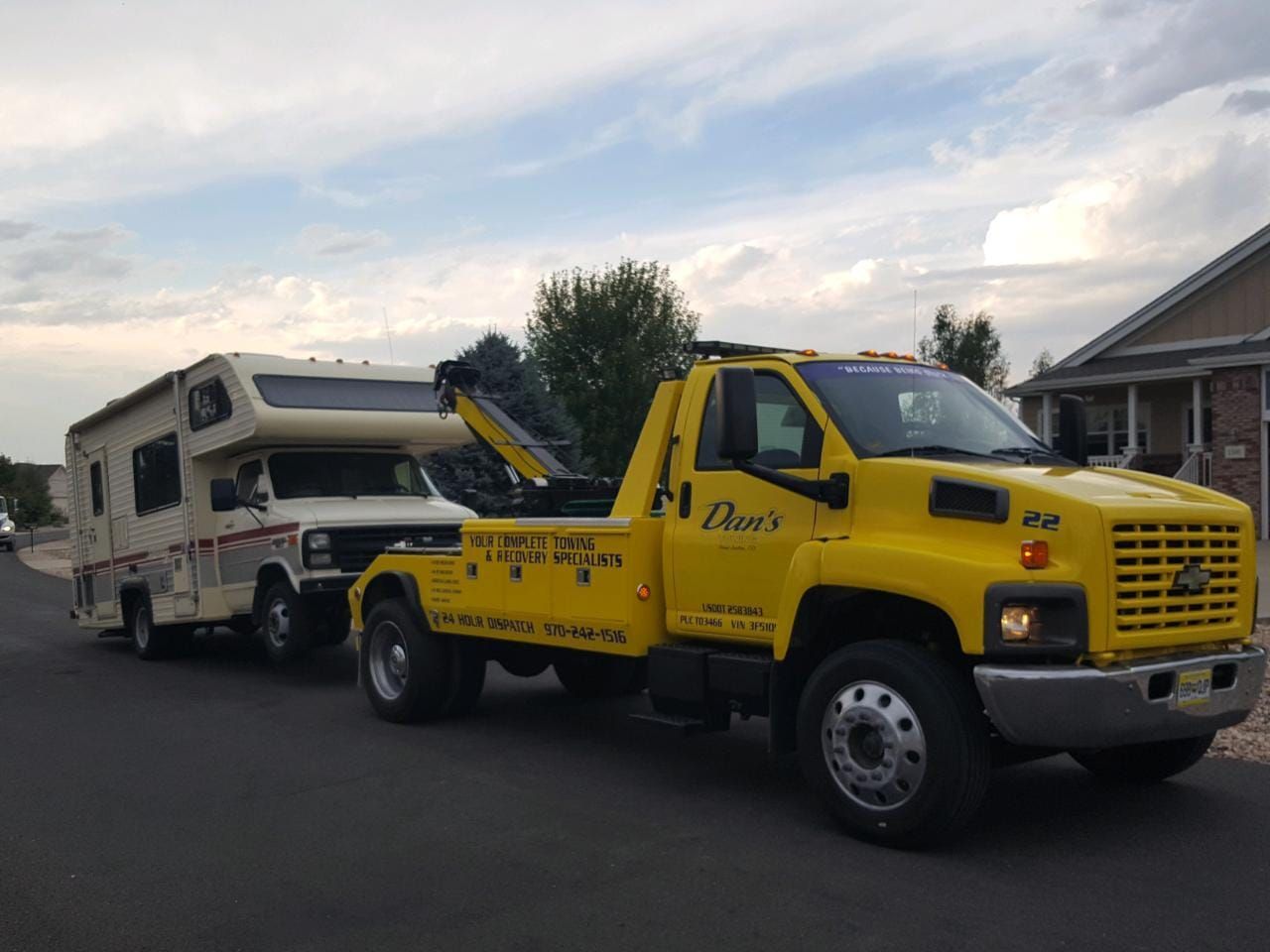 Yellow tow truck towing a beige RV on a suburban street.