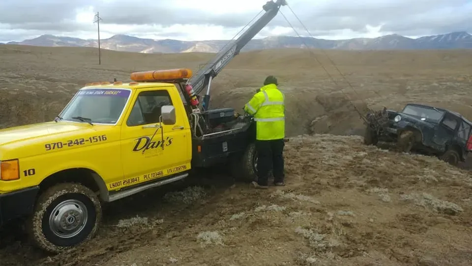 Yellow tow truck pulling a black Jeep from a muddy ditch in a rural setting.
