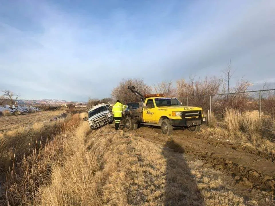Yellow tow truck pulling a white car out of a ditch on a dirt road, under a blue sky.