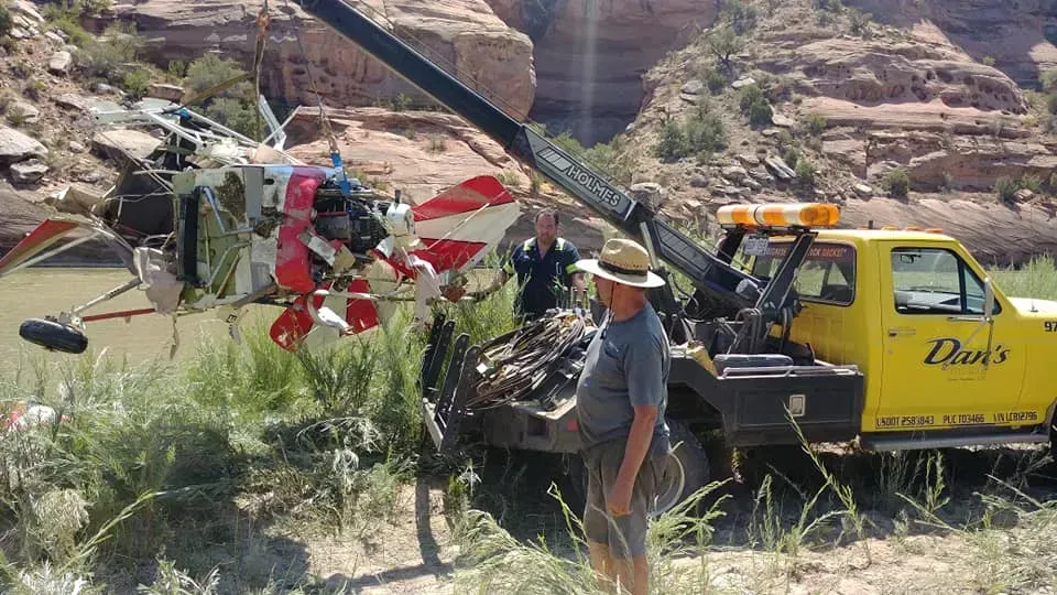 Wrecked helicopter being towed from a riverbed by a yellow tow truck. Men observe near red rock canyon.