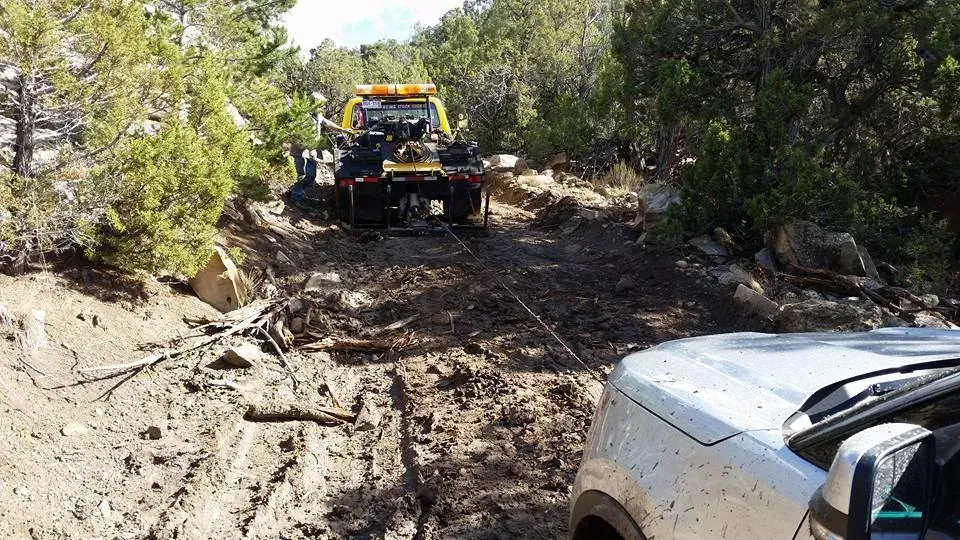 A tow truck pulling a white SUV stuck on a muddy off-road trail.