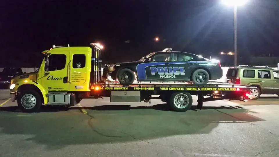 Tow truck carrying a police car at night, with a yellow truck and dark background.