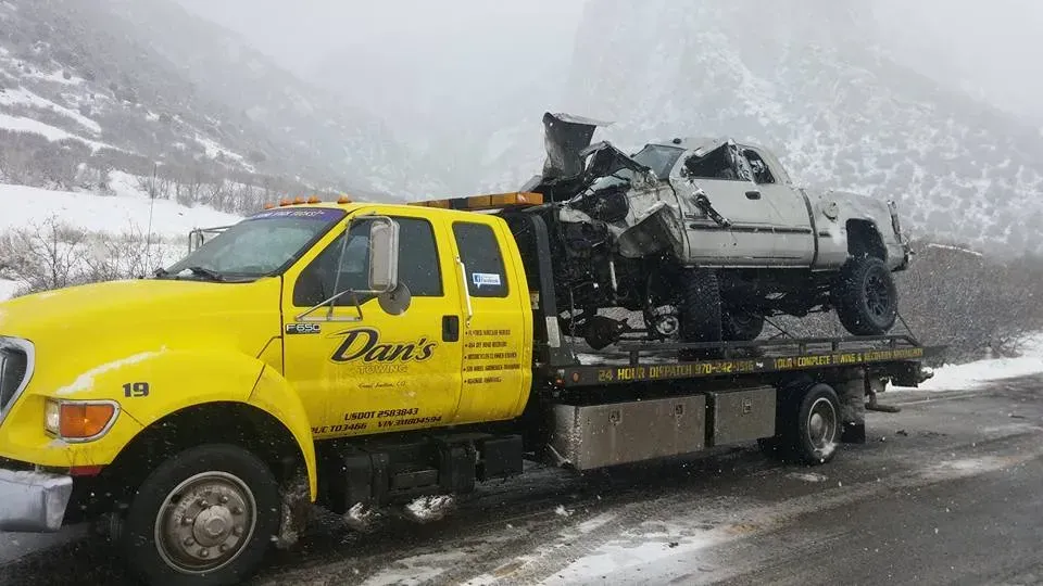 Yellow tow truck hauling a heavily damaged white pickup truck on a snowy road.