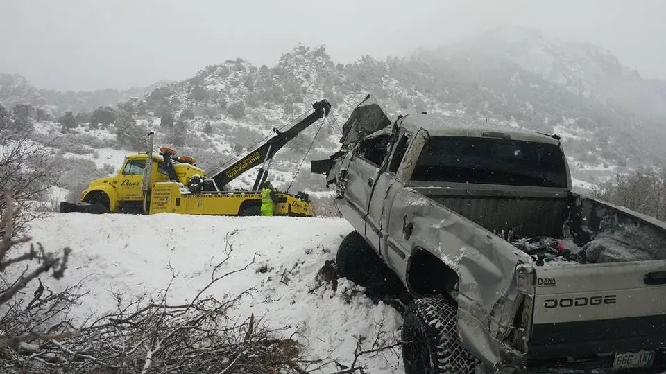 A severely damaged Dodge truck on snowy embankment being towed by a yellow wrecker. Mountains in background.