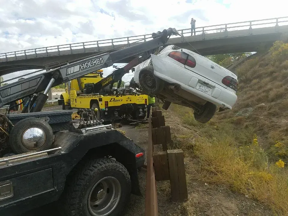 A white car being towed from a ditch by a tow truck next to a bridge.