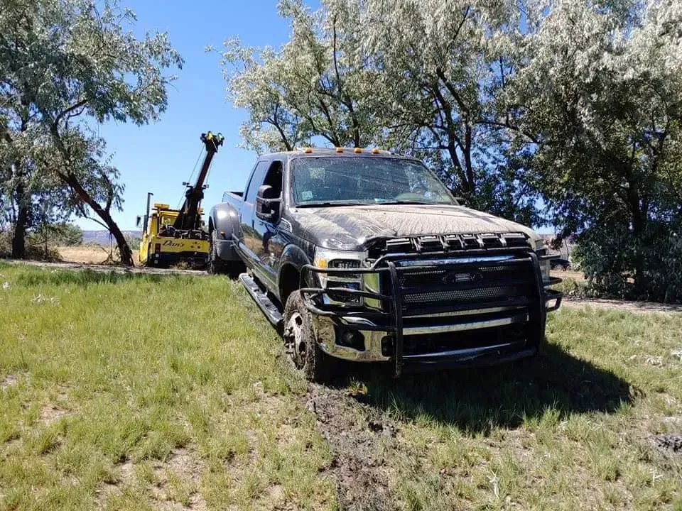 Dark pickup truck being pulled from ditch by a yellow tow truck on a grassy area, under blue sky.