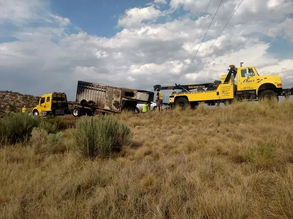 Two yellow tow trucks removing a burned-out semi-truck trailer from a grassy field under a cloudy sky.