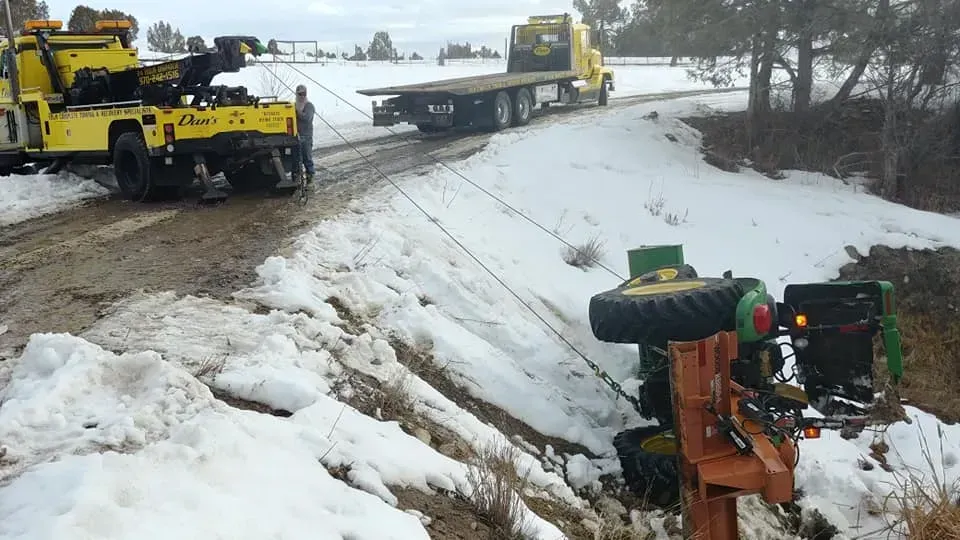 A tow truck attempts to pull a green tractor from a snow-covered ditch on a muddy road.