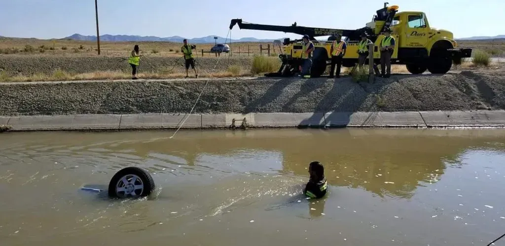 A vehicle submerged in water, being recovered by a tow truck. A person is in the water; others watch from the bank.