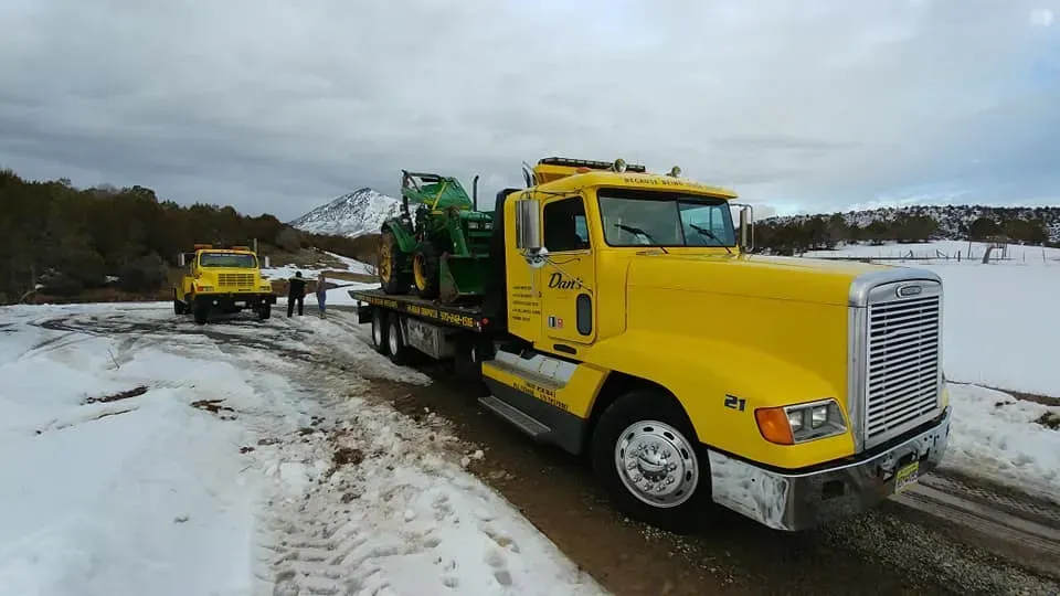 Yellow tow truck with tractor on a snowy road, another truck behind.