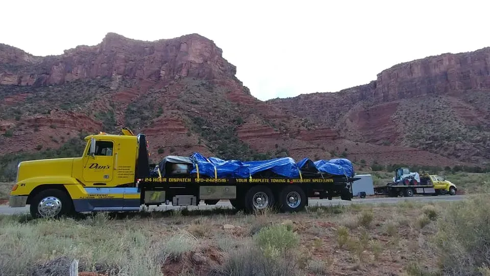 Yellow tow truck on a road with blue-covered cargo and a towed vehicle, mountains in background.