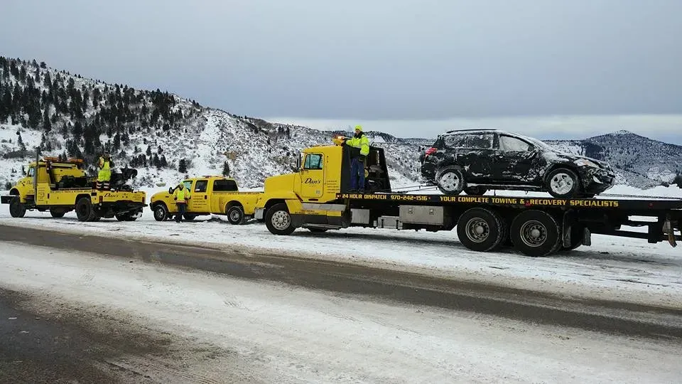 Tow trucks loading a damaged black car on a snowy mountain road.