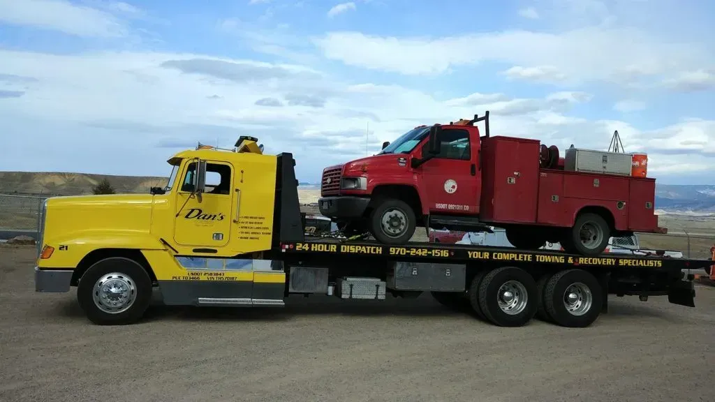 Yellow tow truck carrying a red utility truck on a flatbed, set against a cloudy sky.