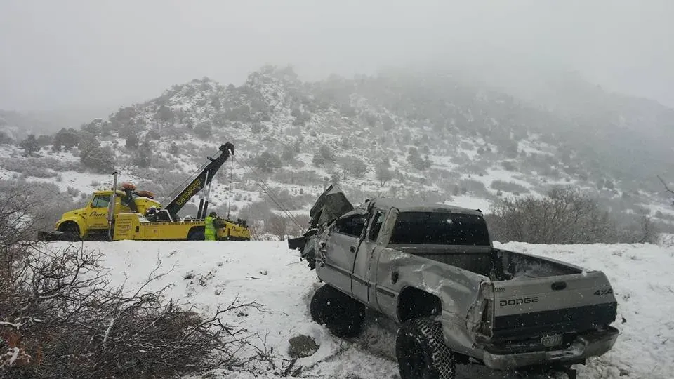 Truck wreckage in snowy landscape being towed by a yellow tow truck.