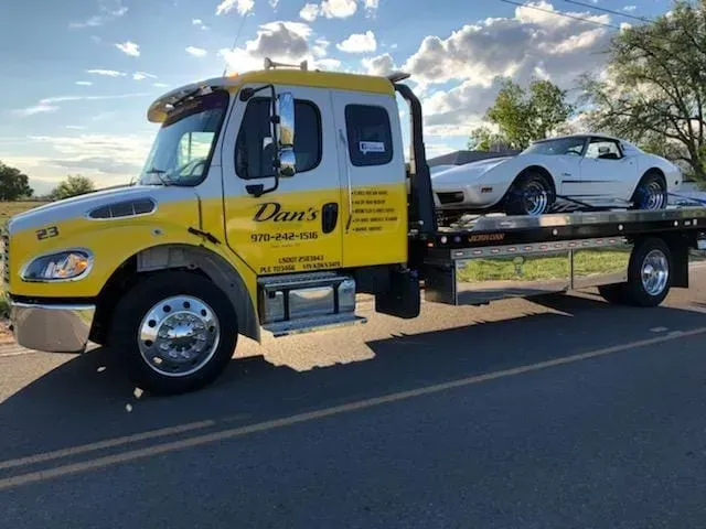 Yellow and white tow truck carrying a white Corvette on a road.