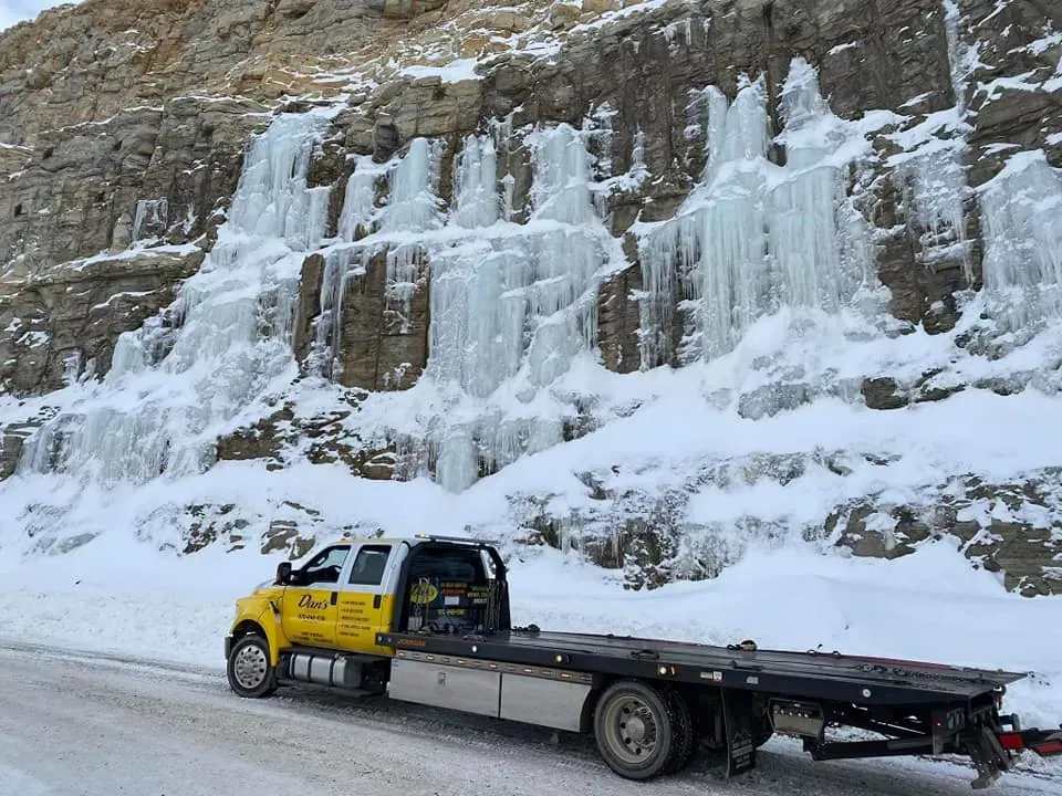 Tow truck on snowy road with frozen waterfalls cascading down a rocky cliff face.