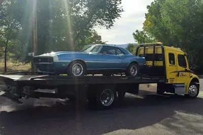 Blue classic car being transported on a yellow tow truck, parked on asphalt.