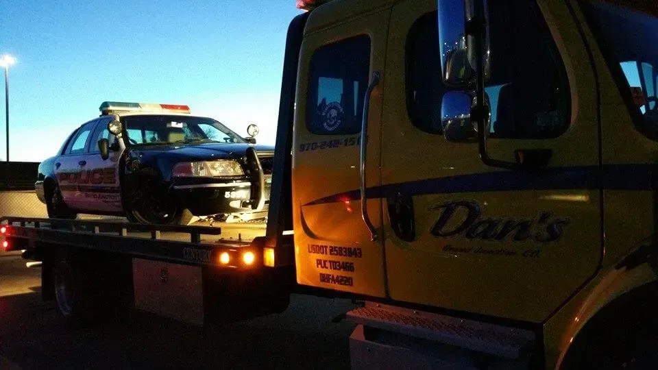 Police car being towed by a yellow Dan's tow truck at dusk.
