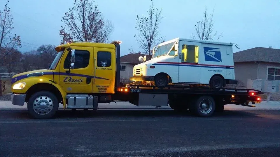 Yellow tow truck carrying a white U.S. Postal Service mail truck on a flatbed, in an evening setting.