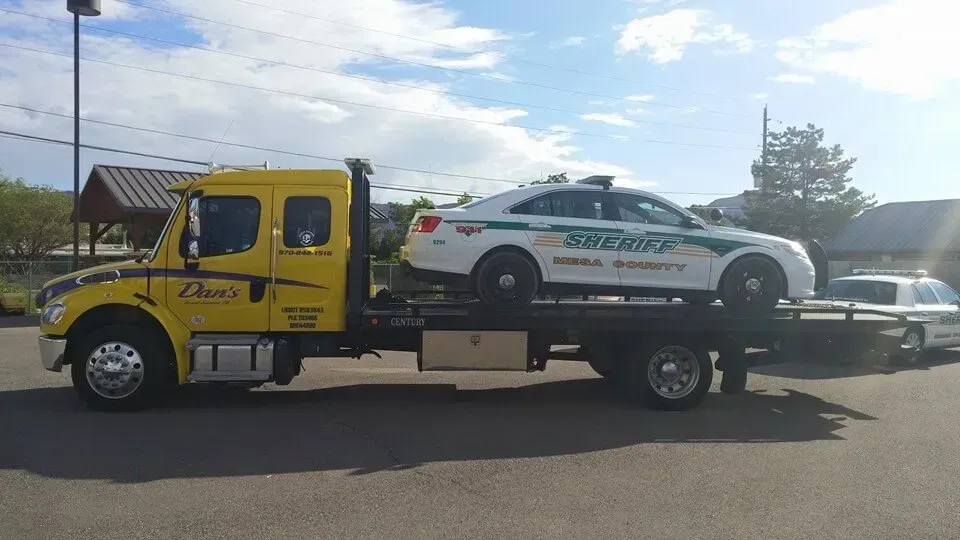 Yellow tow truck carrying a Sheriff's patrol car. Sunny day, parked on pavement.