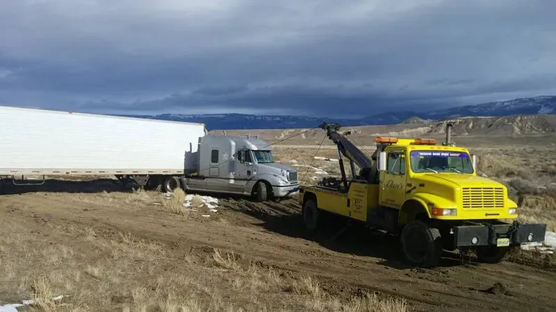 Yellow tow truck pulling a semi-truck out of a muddy ditch on a rural road. Cloudy sky in background.