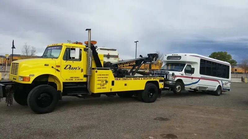 Yellow tow truck towing a white and blue bus in a parking lot under a cloudy sky.