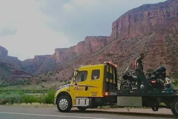 Yellow tow truck with motorcycle on flatbed in front of red rock canyon.