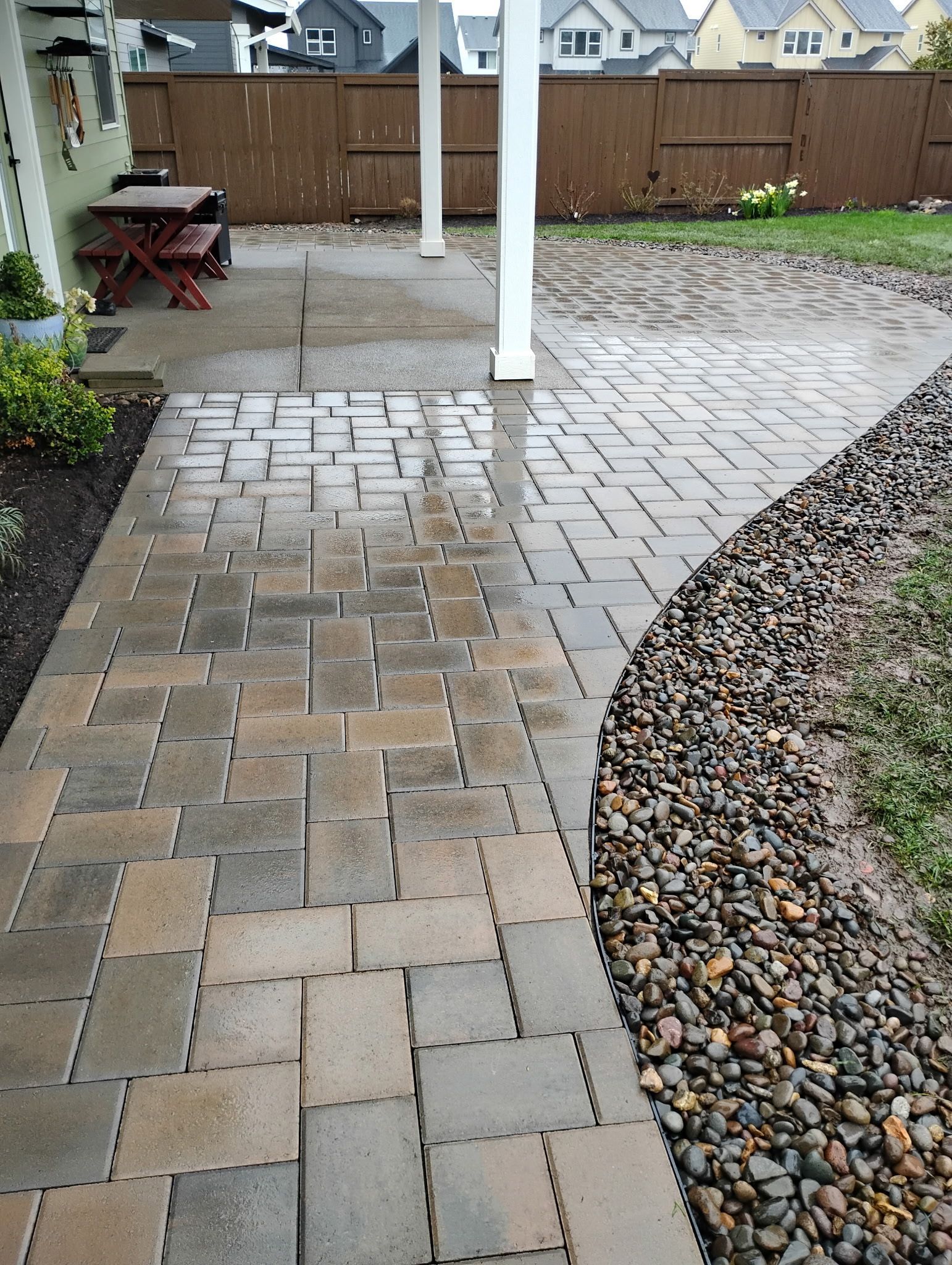 Wet stone patio path beside a gravel border and house siding, with white posts and yard furniture in the background