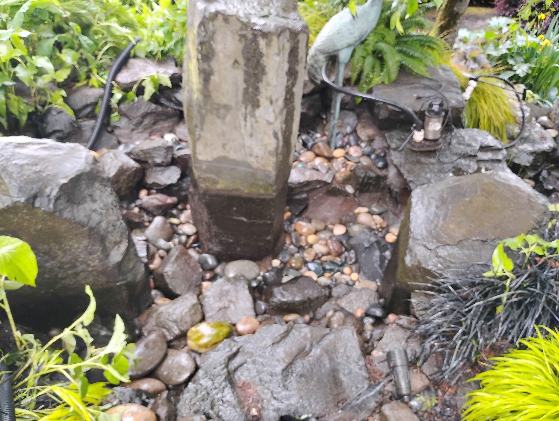 Small garden pond with rocks, a stone pillar, and a heron statue amid green plants