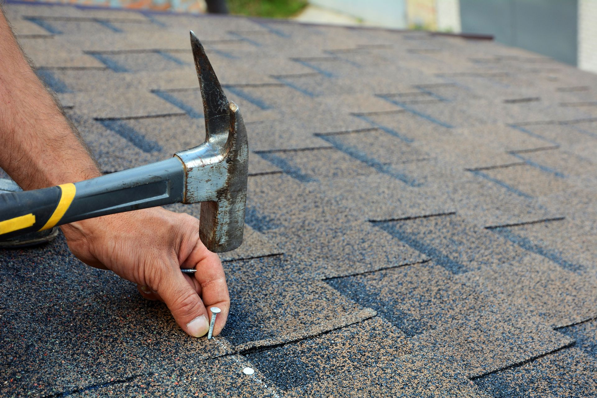 A person’s hand uses a roofing hammer to drive a nail into dark, textured asphalt shingles on a sloped roof.