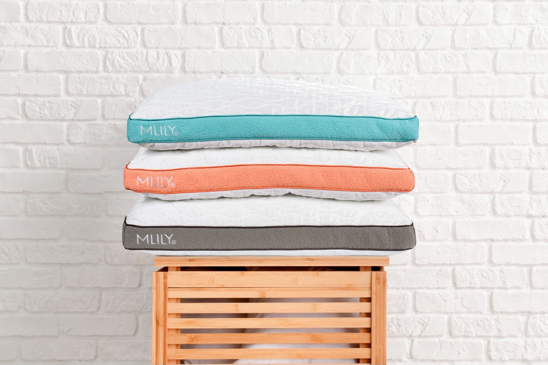 Three colorful pillows stacked on a wooden crate against a white brick wall. Each pillow has a colored trim.