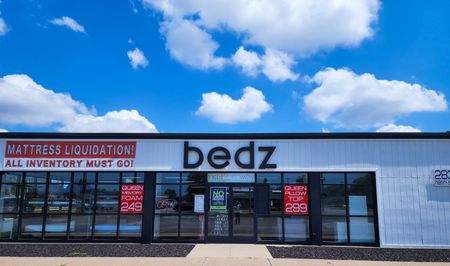 Exterior of "bedz" mattress store with liquidation sale signs, under a blue sky with clouds.