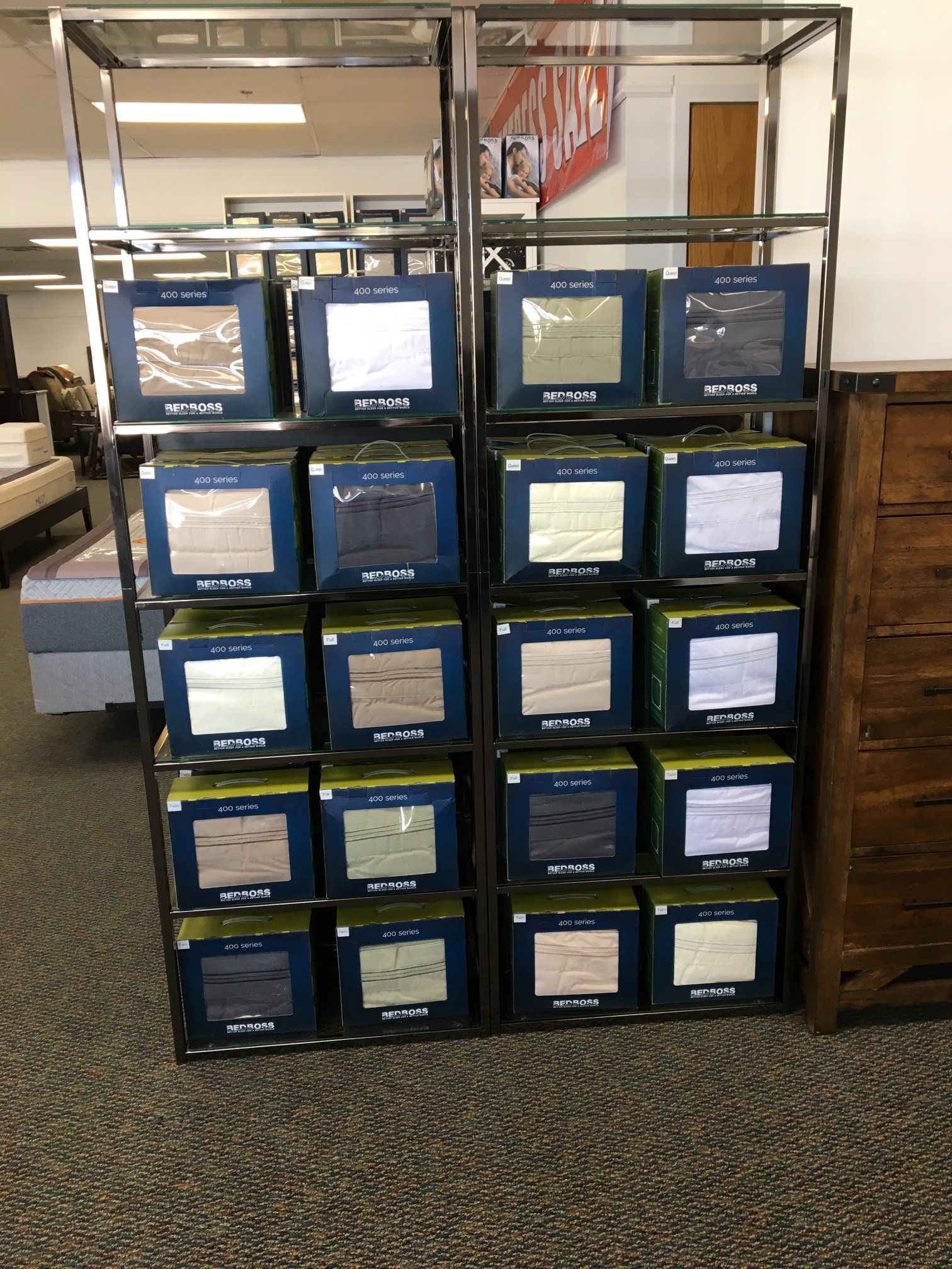 Shelving unit displaying multiple boxed bedding sets in a store, various colors, neutral carpet.
