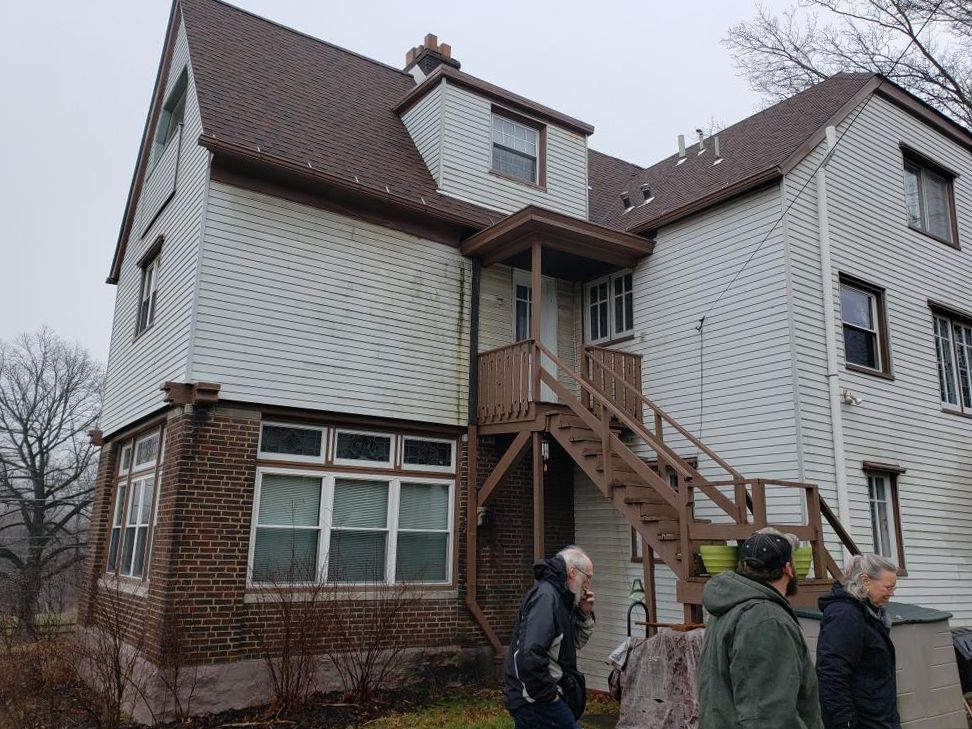 A group of people are standing in front of a house with stairs leading up to the second floor.