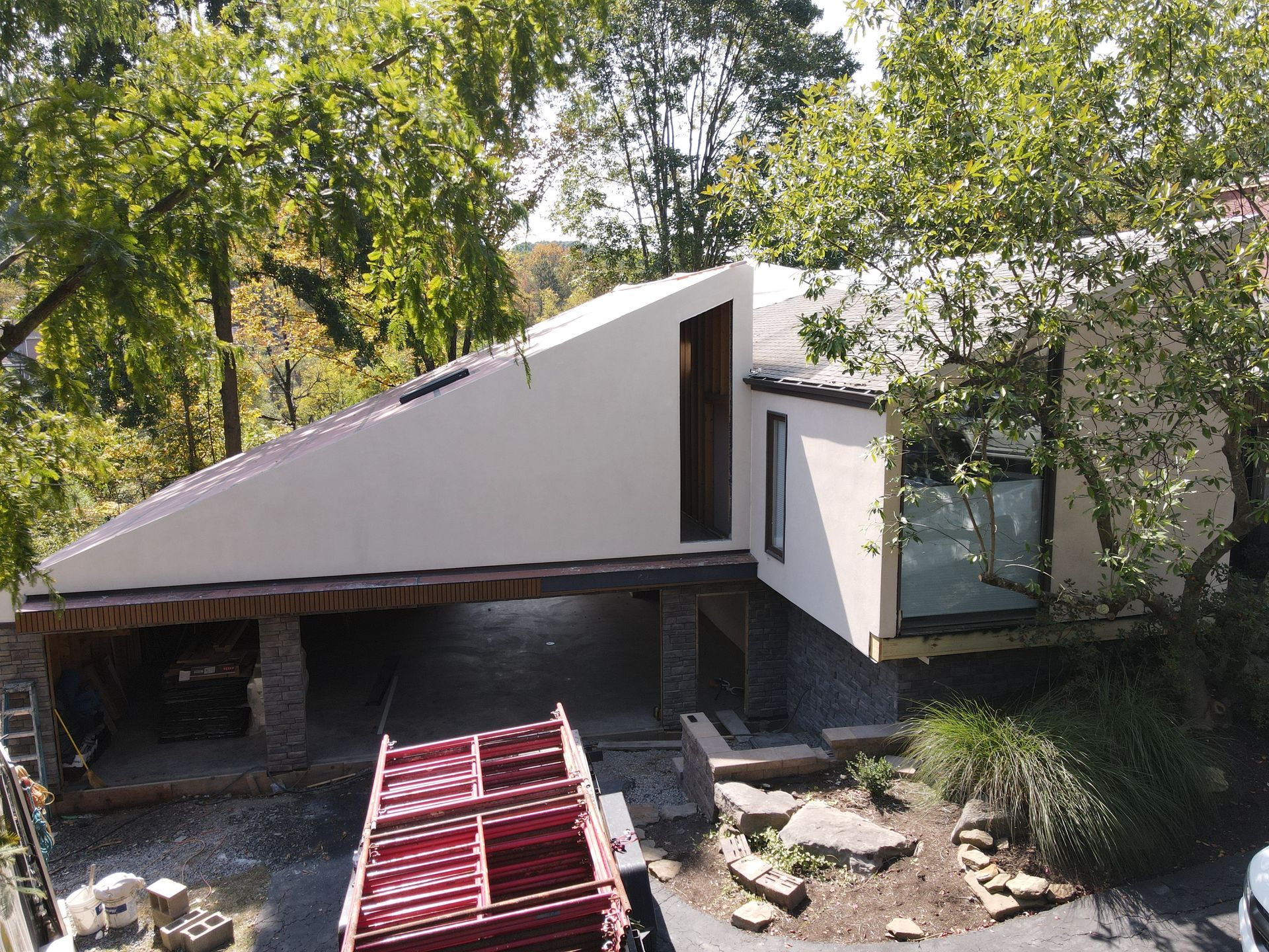 Modern house with angled roof and stone supports, under construction, nestled among trees.