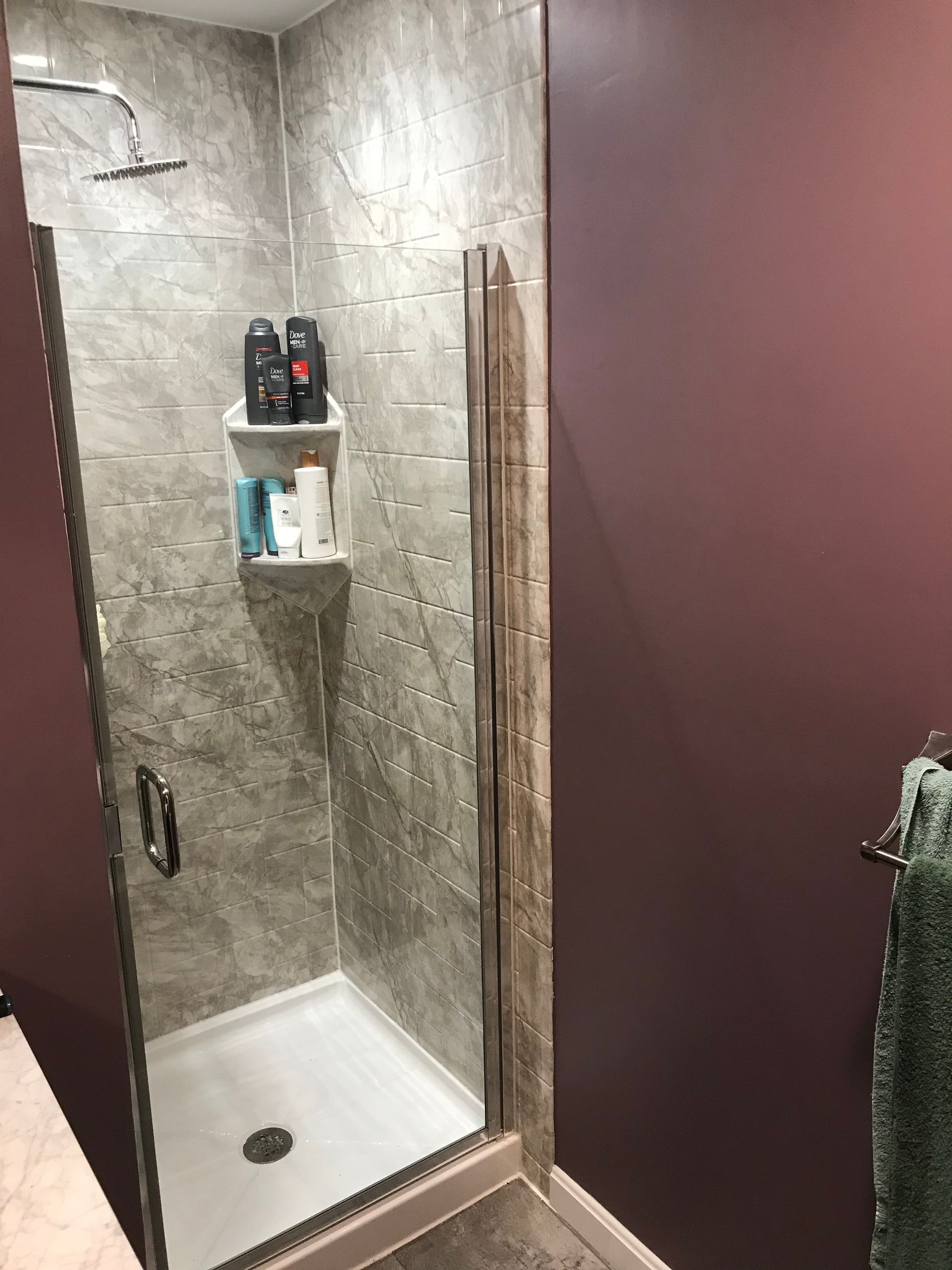 Shower with gray tile, glass door, and a built-in shelf holding toiletries; adjacent to a maroon wall.