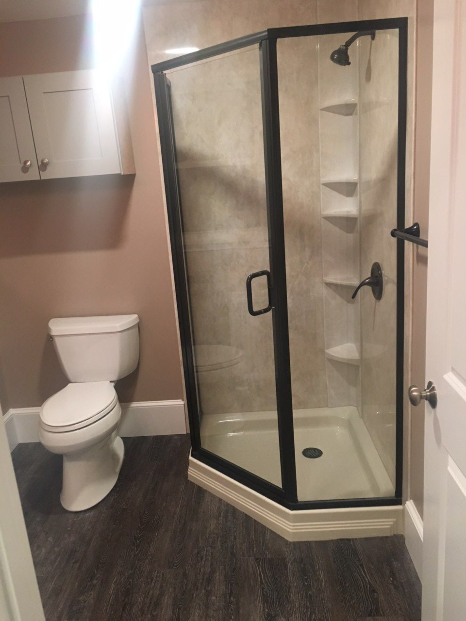 Bathroom with toilet, shower, and cabinet. Gray wood-look floor, light pink walls, and black shower frame.