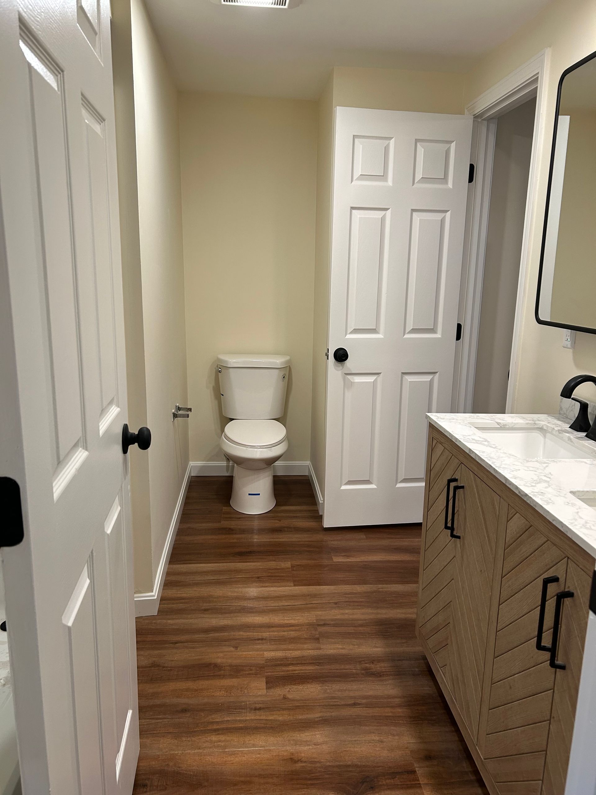 Bathroom with wood-look floor, toilet, vanity, and white door. Light walls, black hardware.